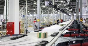 Cardboard packages move along an automated conveyor system inside a large warehouse, with workers and sorting equipment visible in the background.